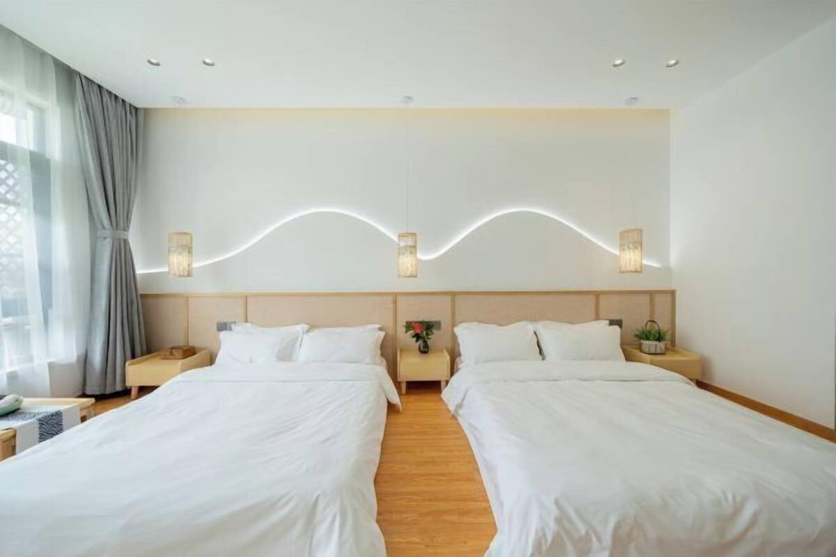 Une chambre d’hôtel avec deux lits, des têtes de lit en bois, des tables de chevet avec des lampes, une grande fenêtre avec des rideaux et un décor mural moderne.