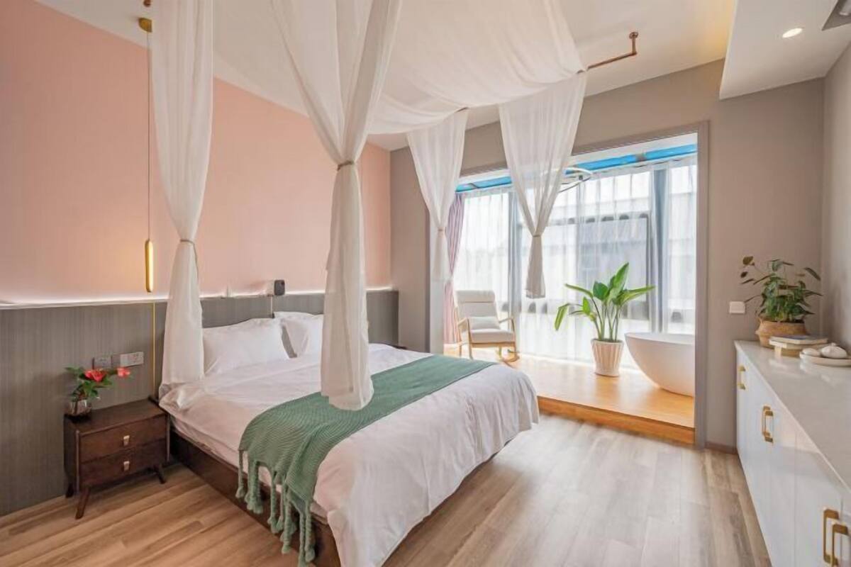 Une chambre moderne avec un lit à baldaquin, une table de chevet avec une lampe, une grande fenêtre offrant une belle vue et une salle de bain avec un lavabo et des plantes.