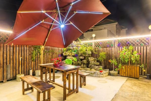 An outdoor patio with a wooden table and benches, a red umbrella, and a wooden fence with plants. - Shanghai Dazaotou Homestay (Shanghai)