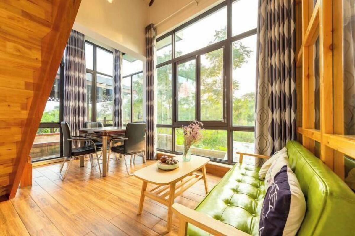 Une pièce dotée d’un plancher en bois, d’un canapé vert, d’une table à manger avec des chaises, et de grandes fenêtres donnant sur un paysage verdoyant.