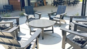 Une terrasse extérieure avec des fauteuils Adirondack et une table ronde, un espace couvert illuminé de guirlandes lumineuses, et une vue sur un champ verdoyant en arrière-plan.