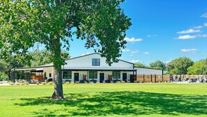 A two-story building with a covered patio area, a large lawn, and a tree in the foreground. - Bungalow 4 @ Barndo Bungalows (Granbury)