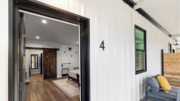 A modern interior with a wooden floor, a bed with a white headboard, a gray sofa with a yellow pillow, and a number '4' on the wall. - Bungalow 4 @ Barndo Bungalows (Granbury)