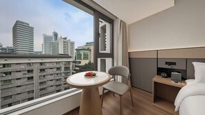 A modern hotel room with a large window, a round table with fruit, a chair, a bed, and a bedside table with a lamp. - Bolianhui Hotel (Chongqing Southwest Hospital) (Chongqing)
