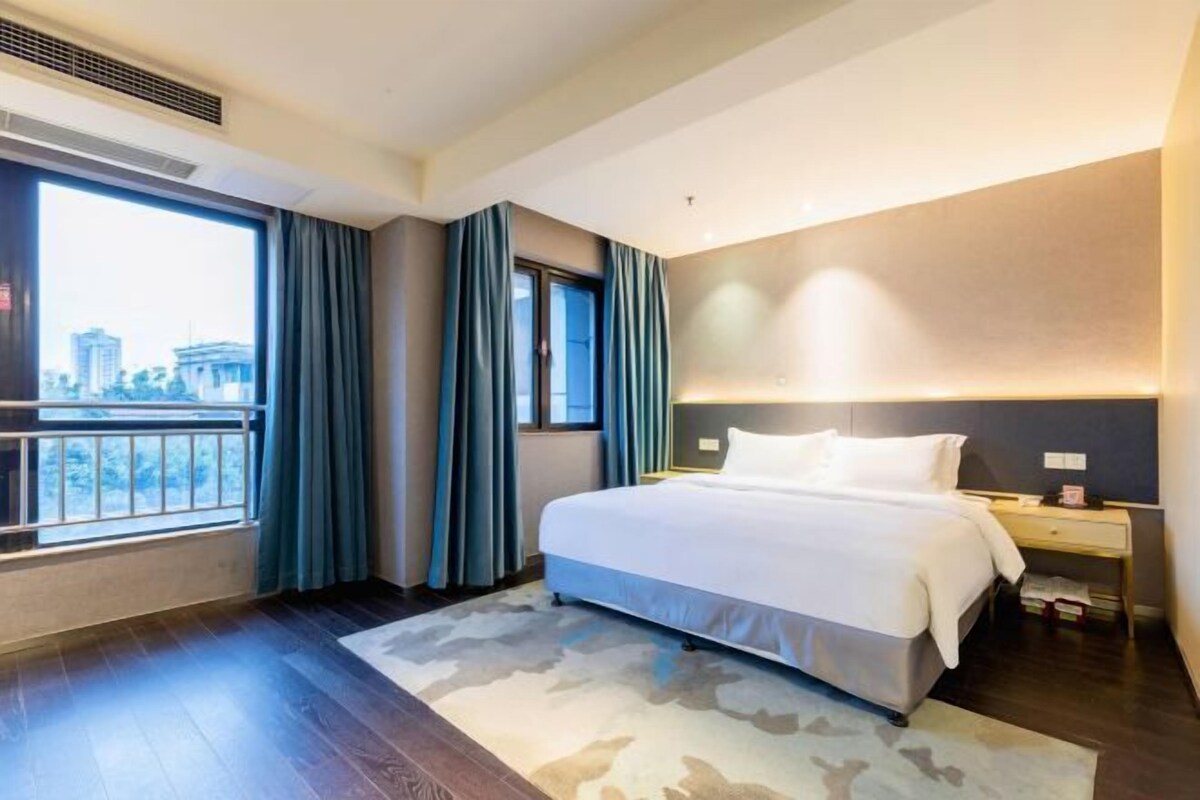 A modern hotel room with a large bed, a nightstand, a window with curtains, and a view of the city.