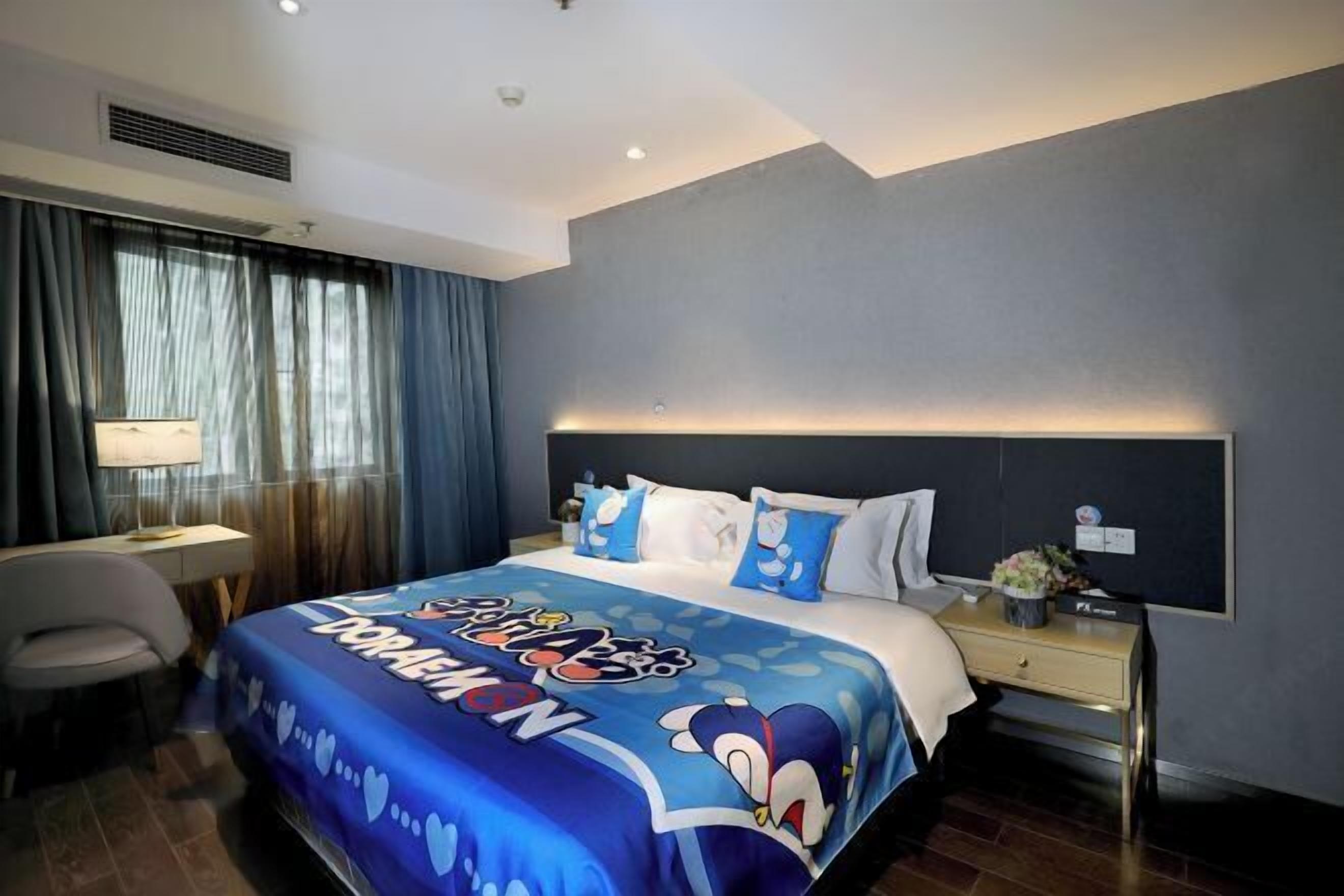 A hotel room with a bed featuring a cartoon character design, a desk with a lamp, and a nightstand with a flower vase.