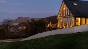 A modern building with large windows and a deck overlooking a body of water and hills. - Sleeps 12, swimming pool, sea views, games rooms (Watermouth)