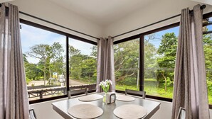 A dining area with a table set for two, large windows, and a view of greenery outside. - Relaxing 2 BR  Beach & Pool  Condo Unit2 (Brasilito)