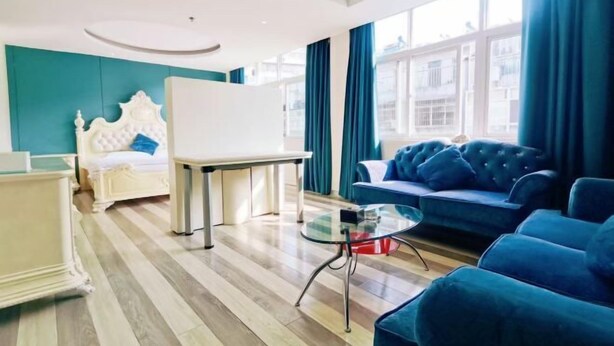 A modern hotel room with a bed, a glass coffee table, a blue sofa, and a view of the city through large windows. - Margarita Boutique Hotel (Guangde)