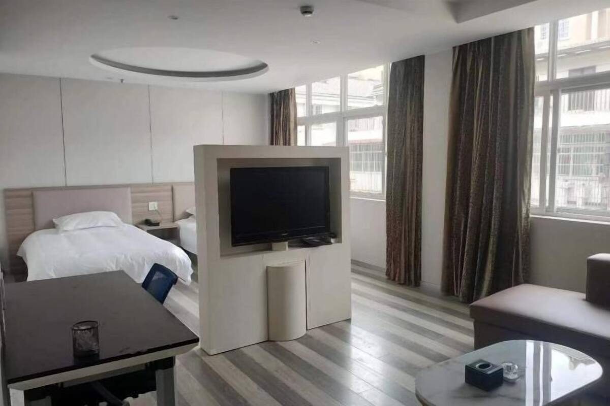 Habitación de hotel con cama, televisión, mesa, sofá y ventana con cortinas.
