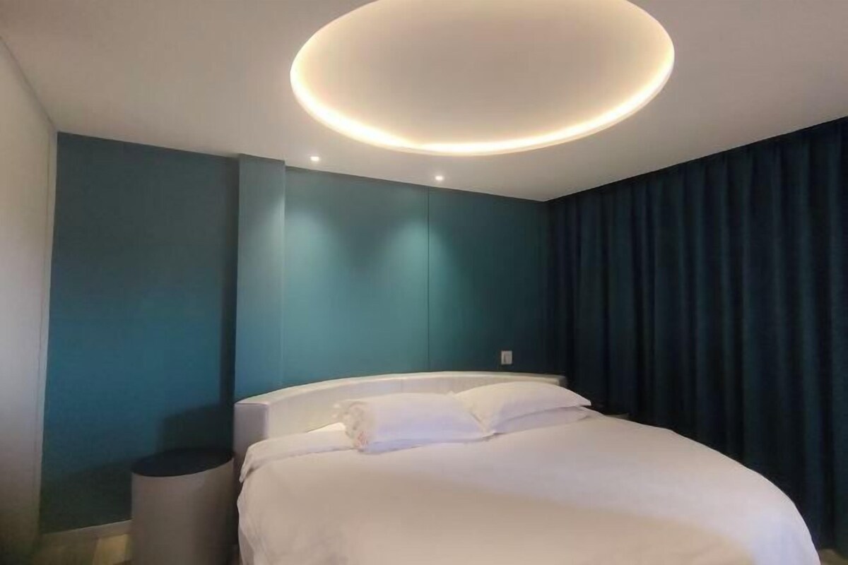 Quarto de hotel moderno com cama, luminária circular no teto, paredes verde-água e cortina escura.