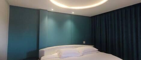 Una habitación de hotel moderna con una cama, una lámpara de techo circular, paredes color turquesa y una cortina oscura.