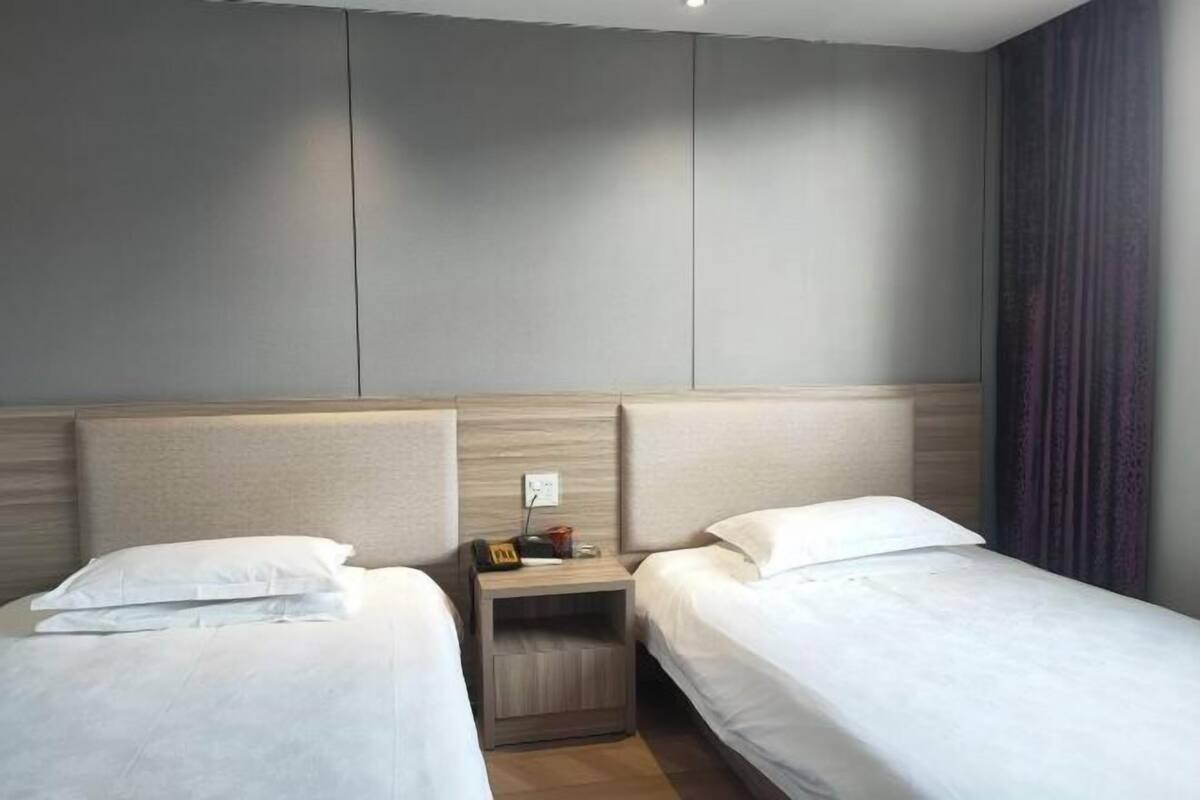 Habitación de hotel con dos camas individuales, una mesita de noche con lámpara y una pared lisa.
