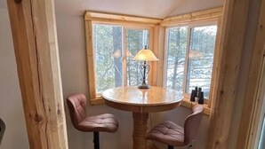 Een hoekkamer met een houten tafel en twee leren barstoelen, een raam met uitzicht op bomen, en een lamp op de tafel.