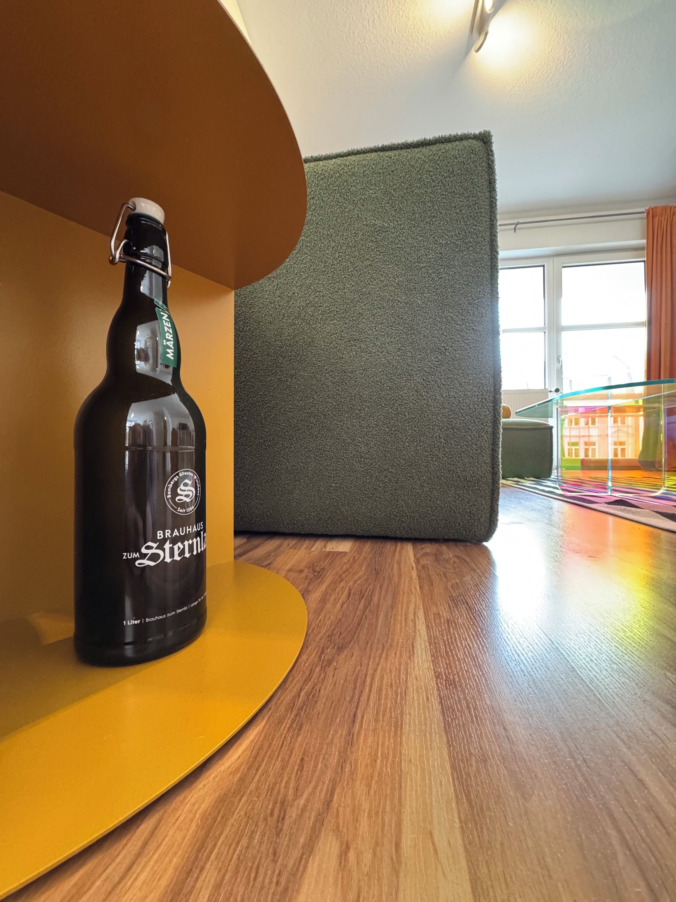 A bottle of beer on a wooden table with a green textured wall and a window in the background.