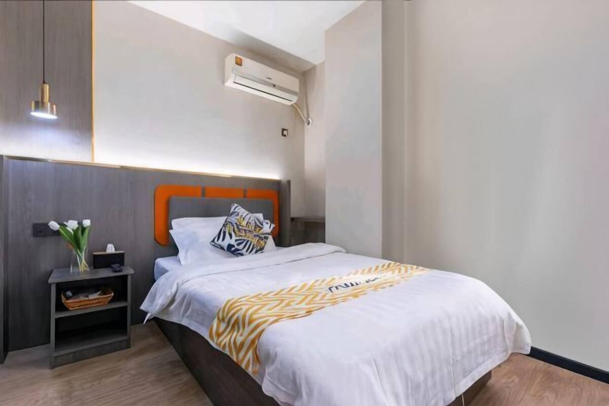 A hotel room with a bed, a nightstand, a vase with flowers, and an air conditioning unit.