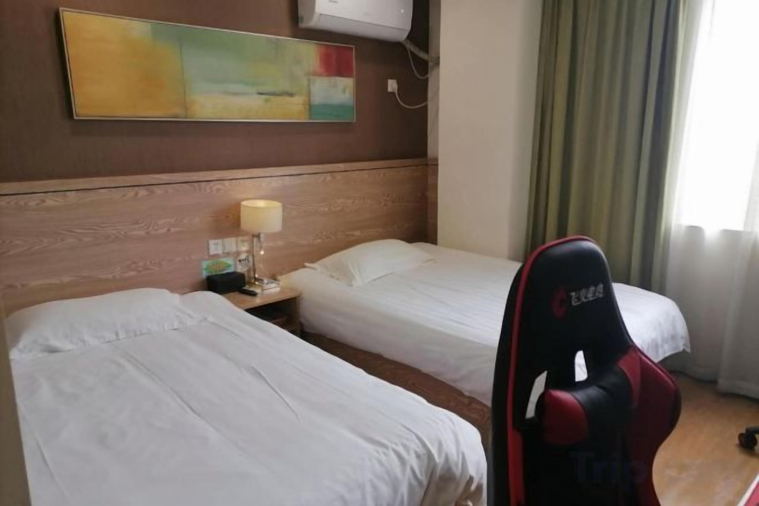 Habitación de hotel con dos camas, un escritorio con lámpara, una silla gamer roja y un cuadro en la pared.