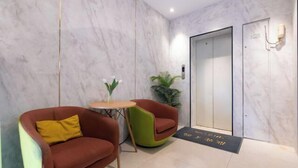 Un hall d’entrée moderne avec un mur de marbre, un comptoir d’accueil, deux fauteuils, une petite table avec un vase et une porte avec une pancarte.