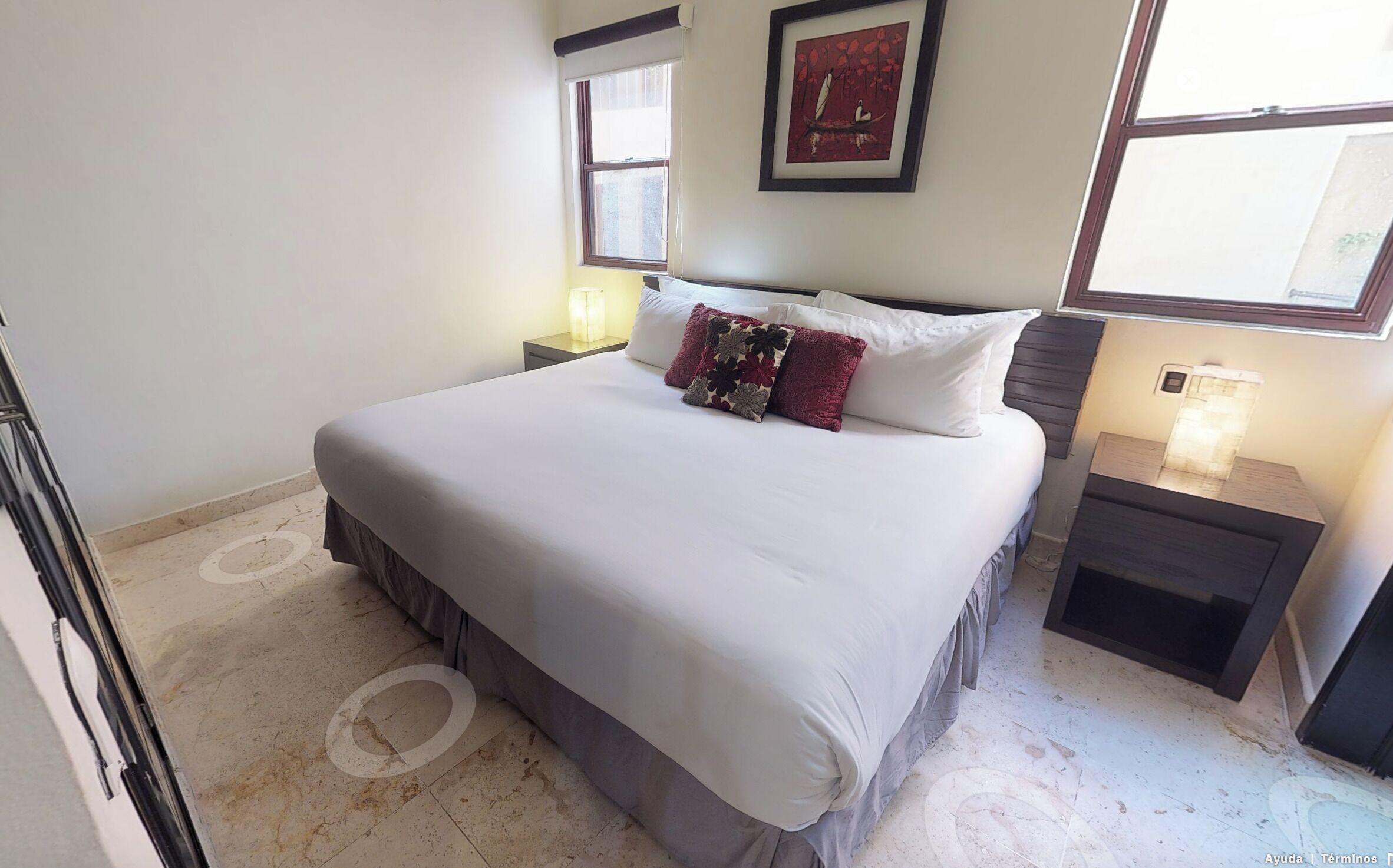 A neatly made bed with a white comforter and a floral pillow, a bedside table with a lamp, and a framed picture on the wall.