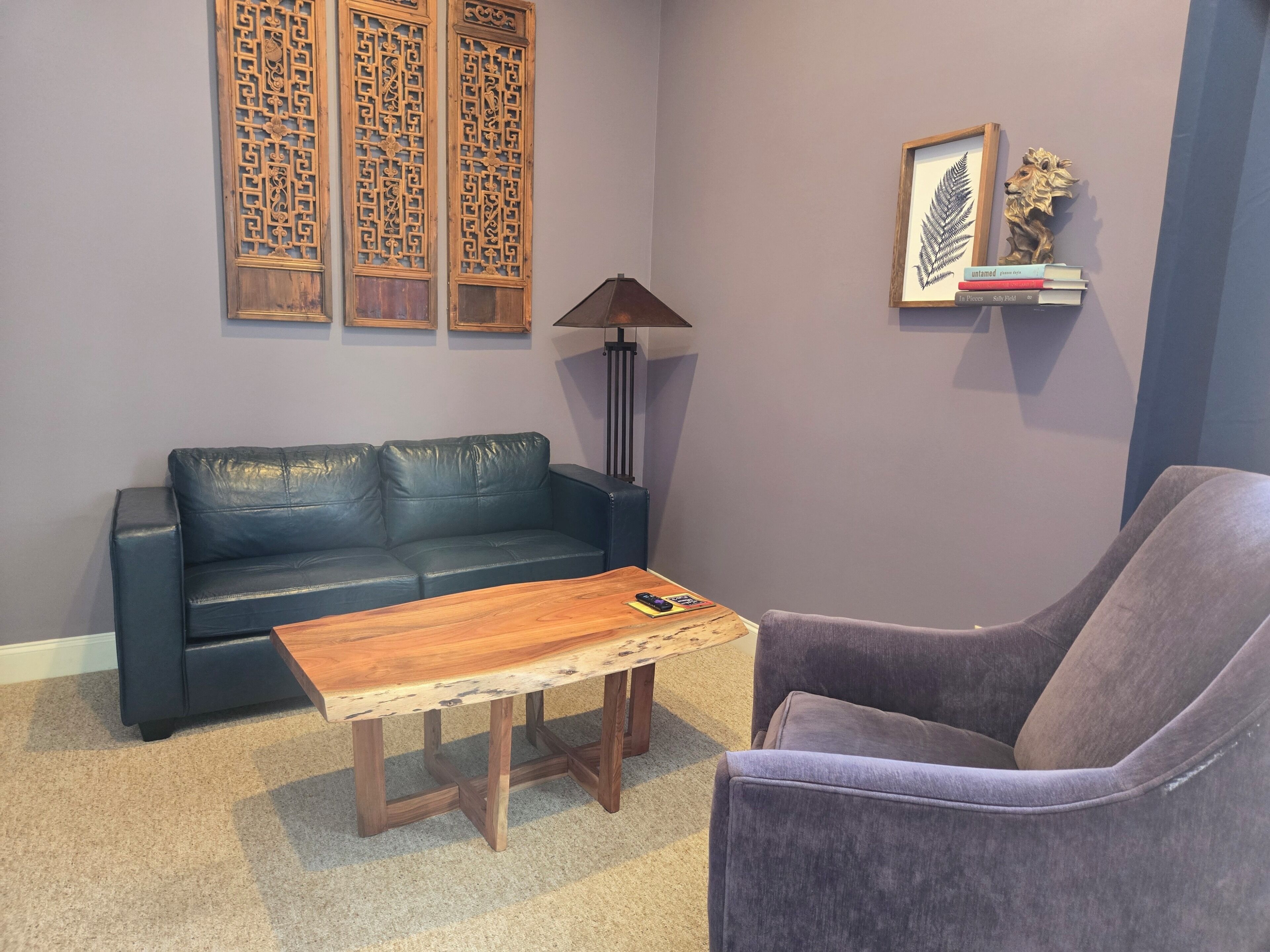 A living room with a dark blue leather sofa, a wooden coffee table, a purple armchair, a floor lamp, and decorative wall panels.