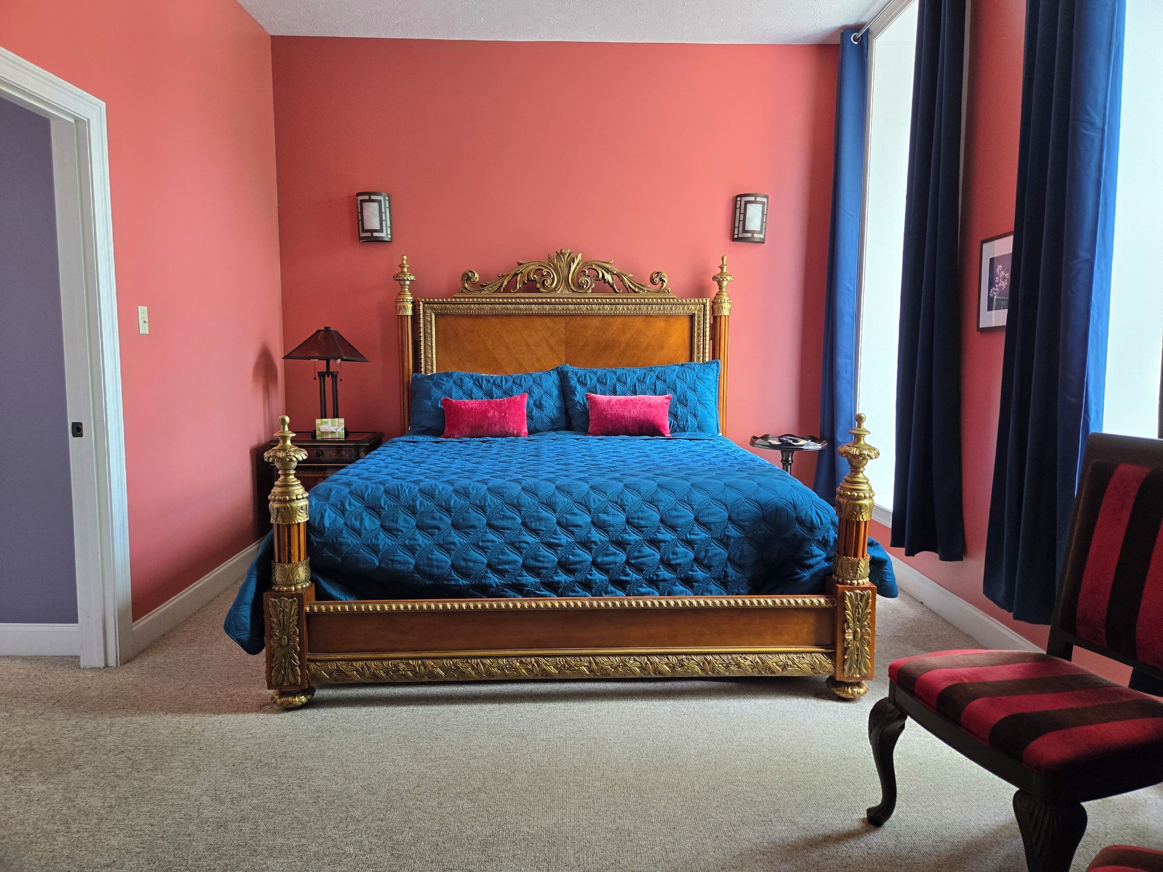 Quarto com cama grande, mesa de cabeceira, cadeira e janela com cortinas.