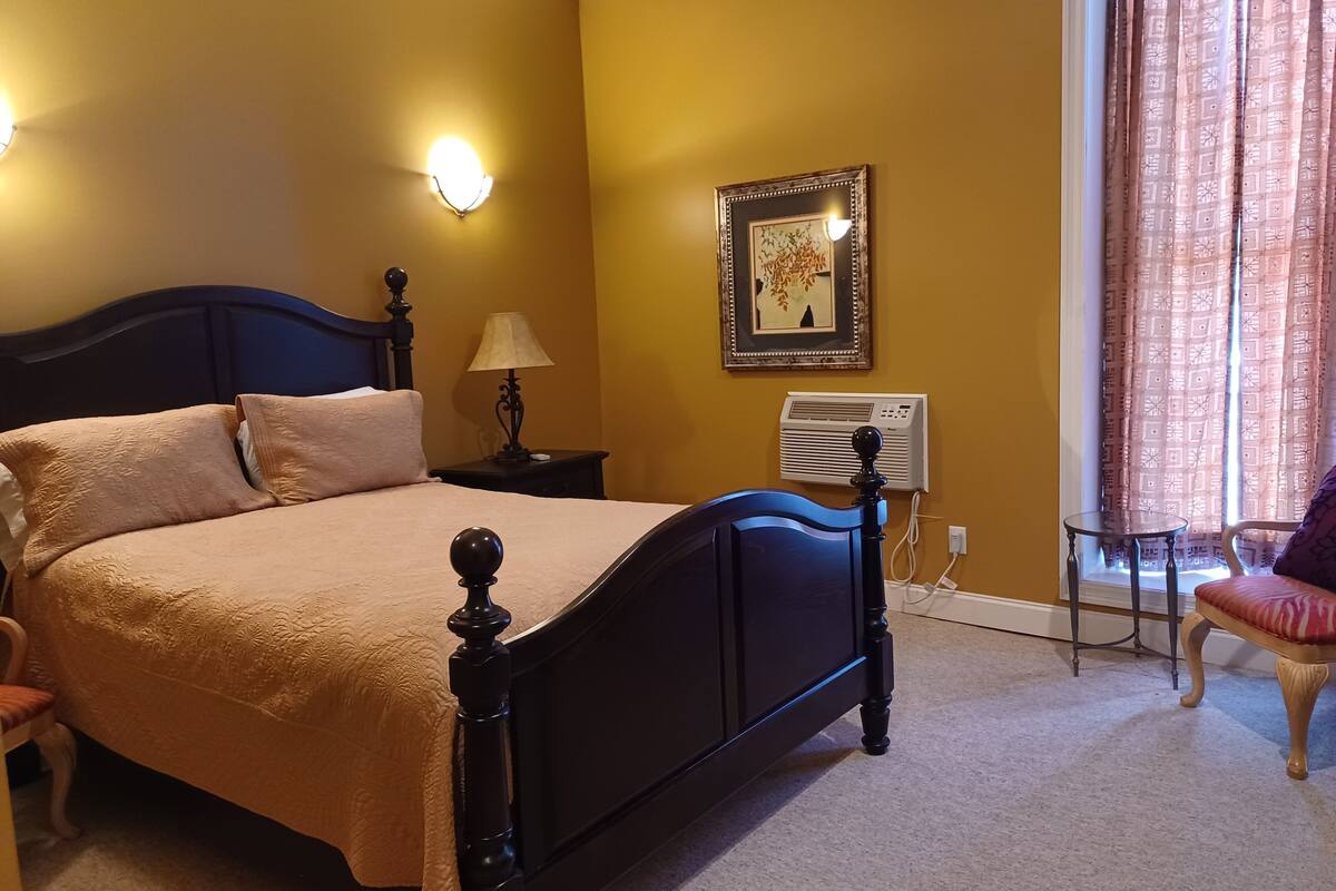 A bedroom with a bed, bedside lamp, air conditioning unit, and a window with curtains.