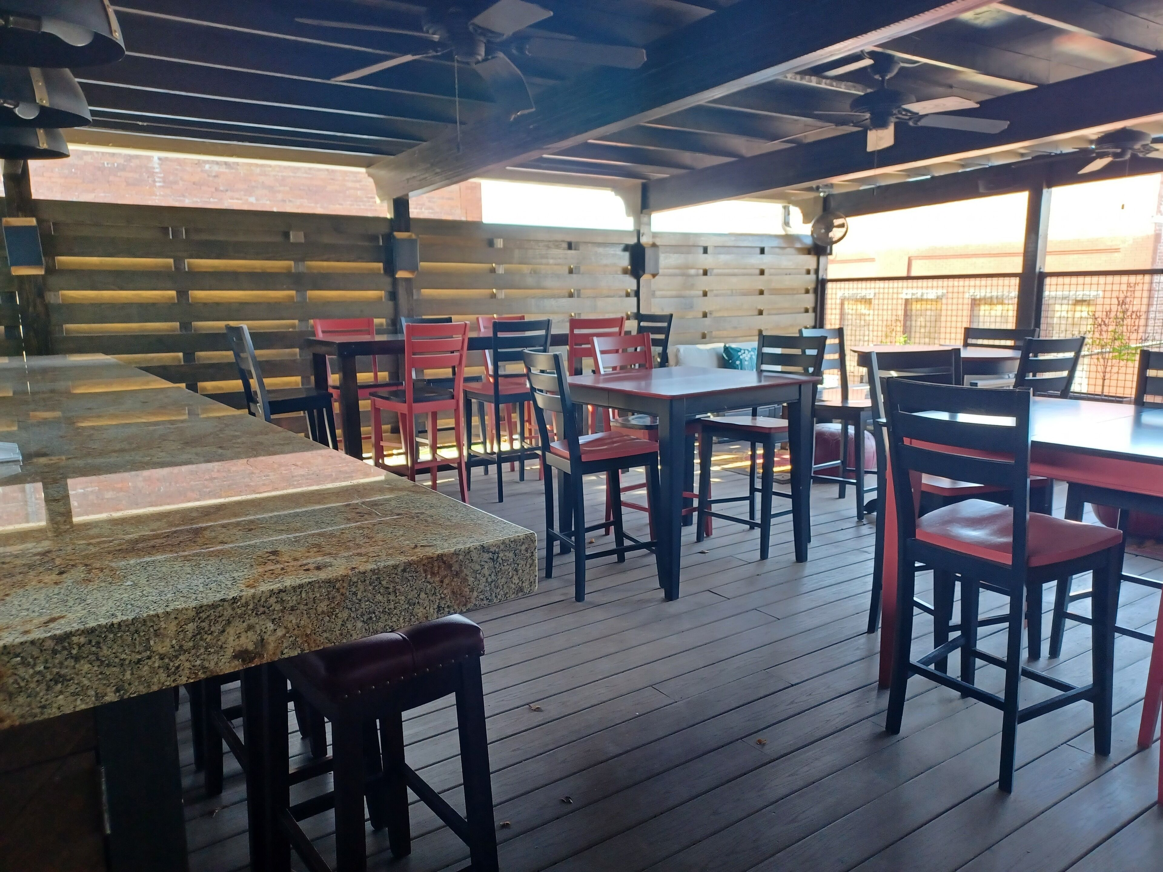 Un patio al aire libre con mesas y sillas de madera, un ventilador de techo y una pared de ladrillo al fondo.