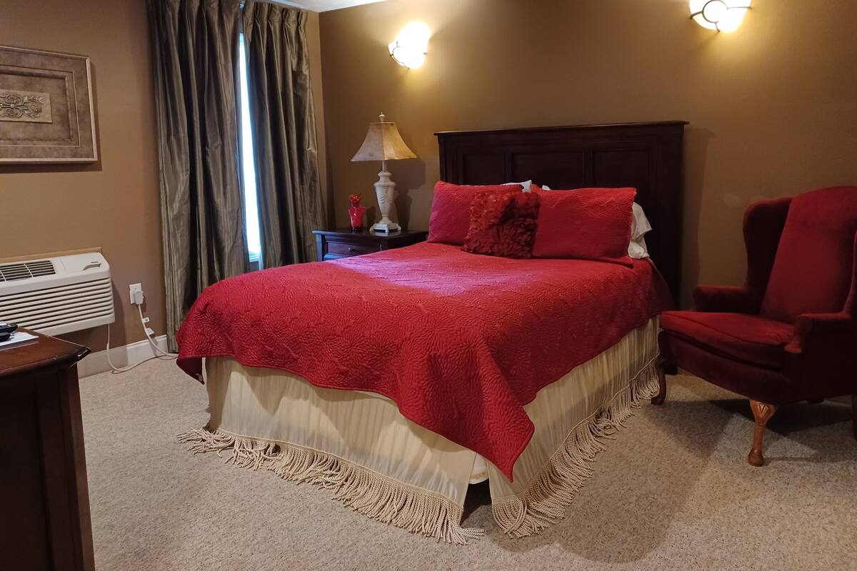 A hotel room with a large bed, a red chair, a nightstand with a lamp, and a wall-mounted light fixture.