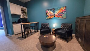 A living room with a coffee table, two armchairs, a painting on the wall, a kitchen area with white cabinets, and a wooden table. - Natchez Manor Bed & Breakfast (Natchez)