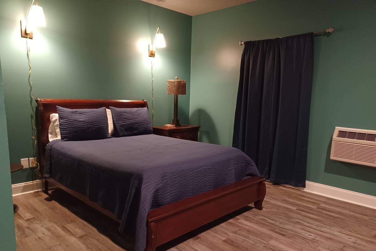 A bedroom with a wooden bed, two bedside lamps, a nightstand, a window with a dark curtain, and a wall-mounted air conditioner.