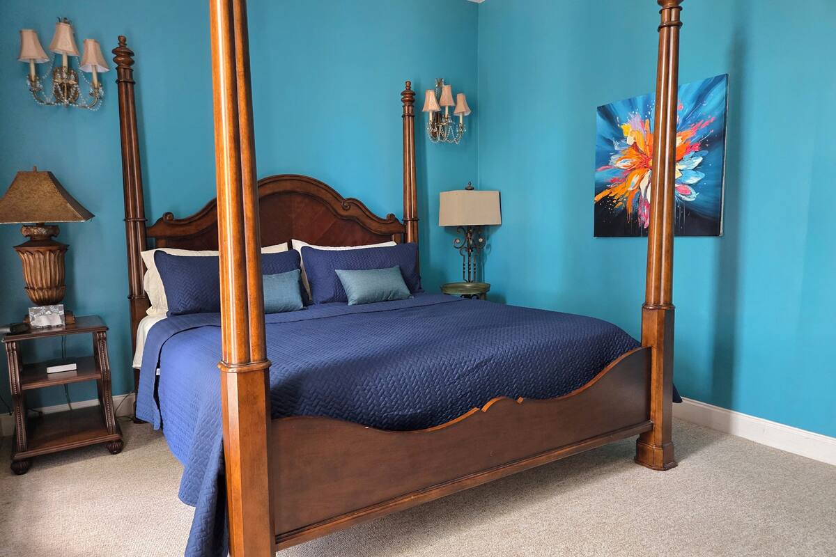 A four-poster bed with a dark wooden frame, a blue quilted bedspread, and matching pillows. There are two bedside tables with lamps, a wall sconce, and a painting.