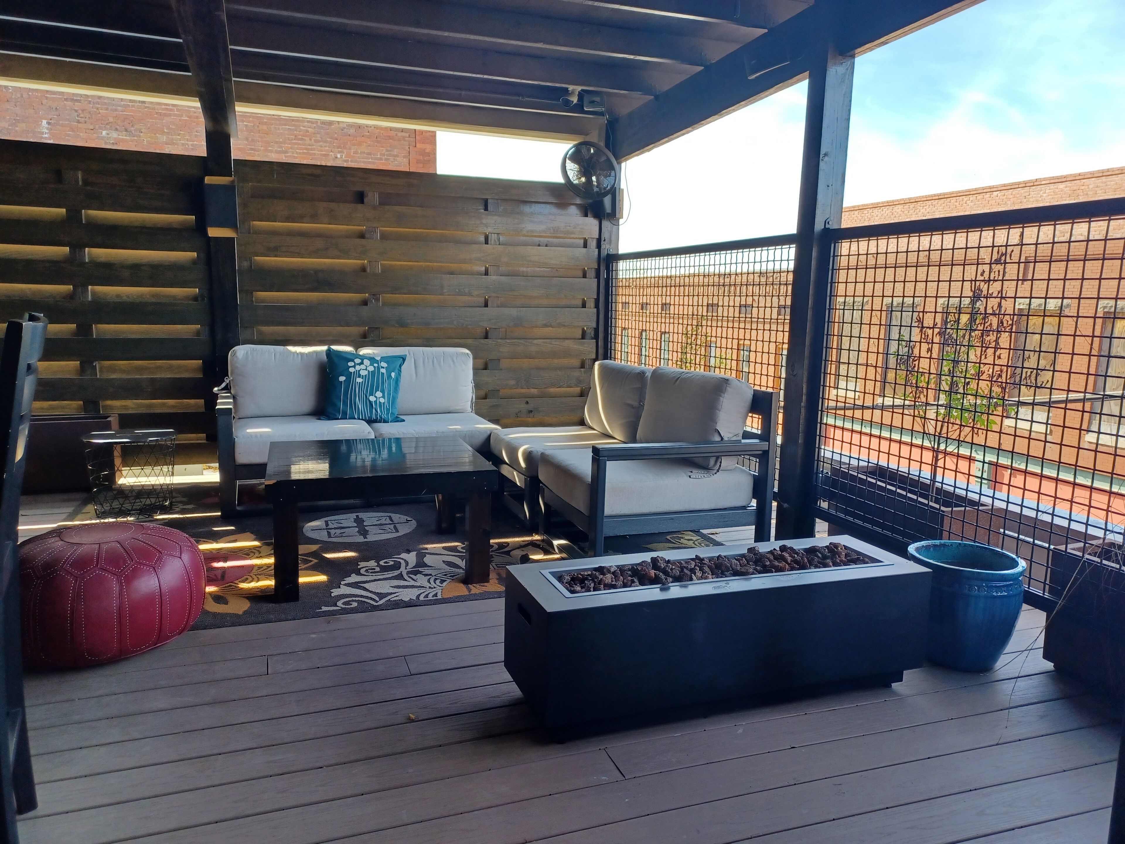A covered outdoor seating area with a wooden floor, a sofa, a coffee table, and a fire pit.