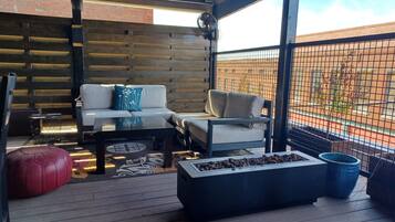 A covered outdoor seating area with a wooden floor, a sofa, a coffee table, and a fire pit.