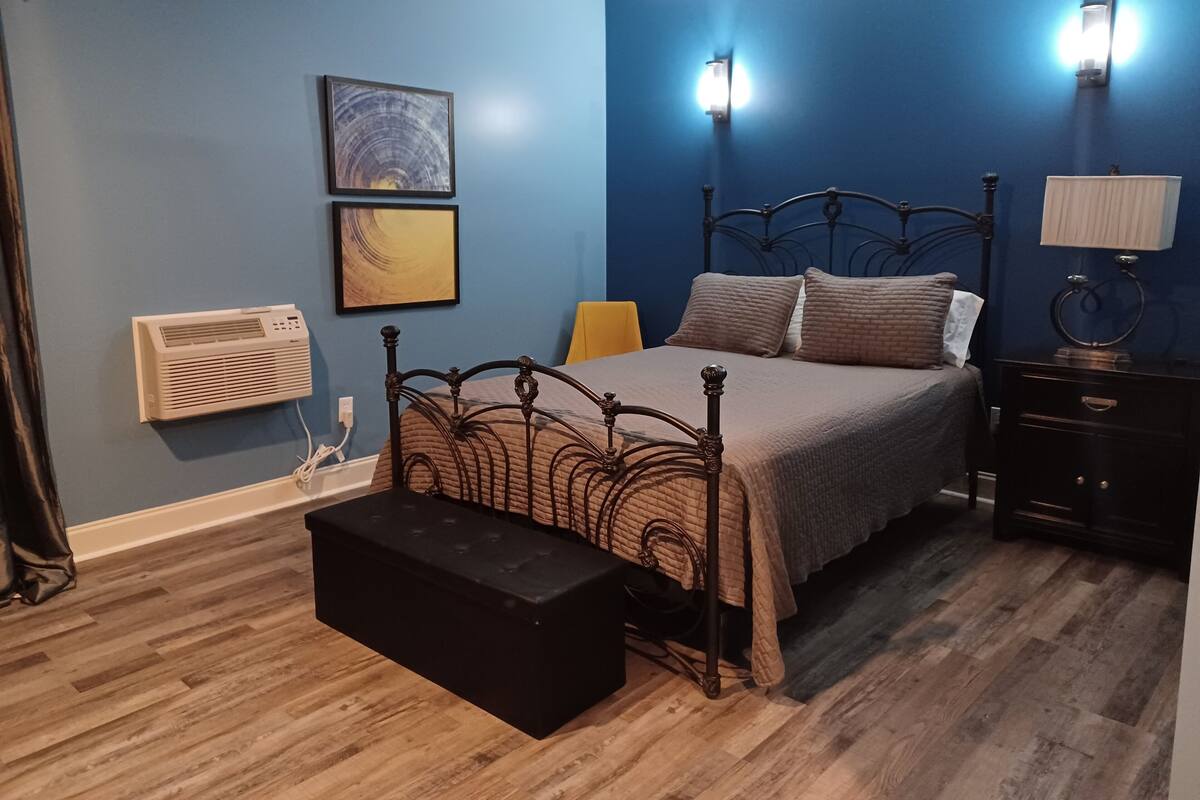 A bedroom with a bed, bedside tables, a nightstand, a wall-mounted air conditioner, and a framed picture on the wall.