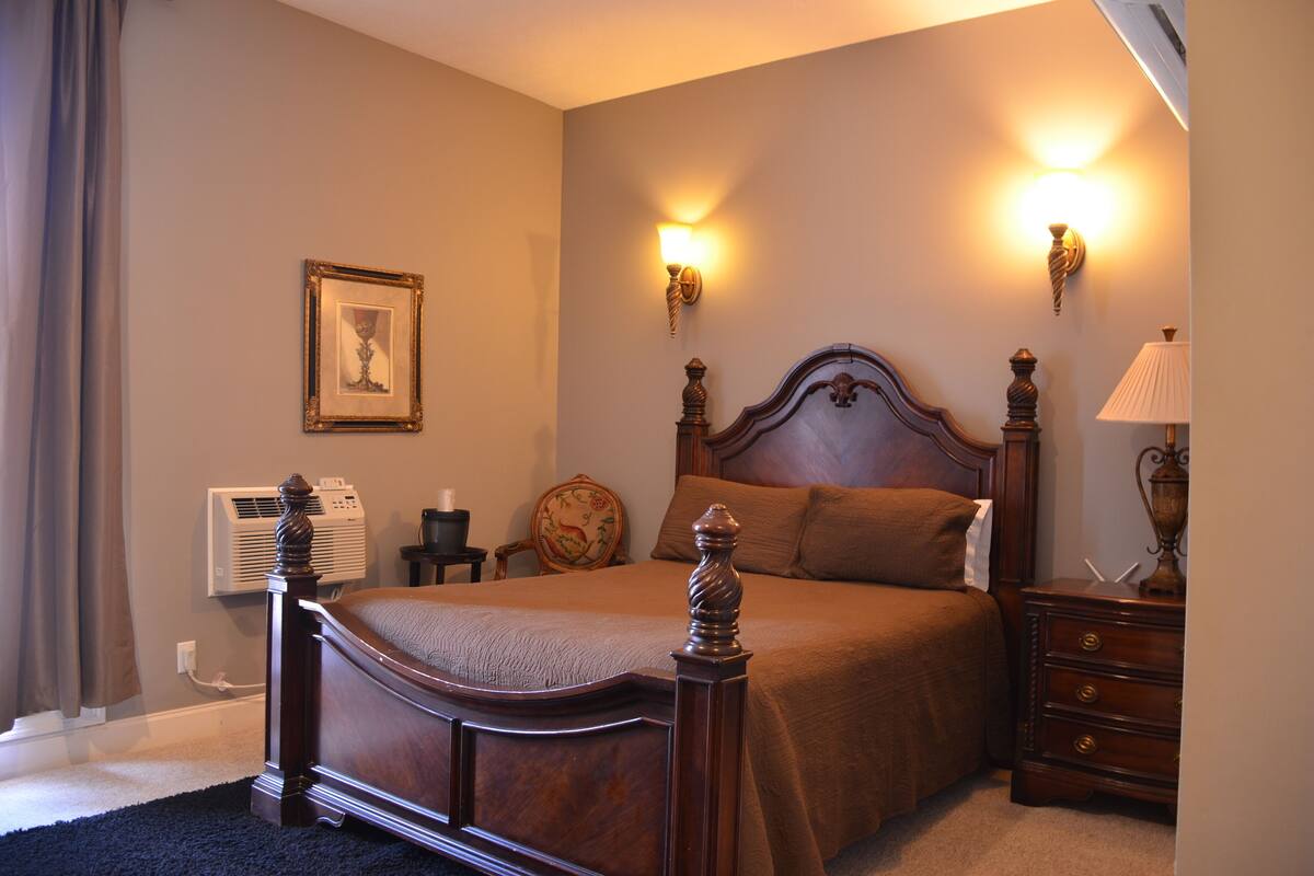 A bedroom with a wooden bed, two wall-mounted lamps, a bedside table, a nightstand, a framed picture, and an air conditioning unit.