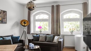 A modern living room with a sofa, coffee table, and floor lamp. There are two windows with curtains, a radiator, and a television. - Old town! Exclusive 3-room apartment, roof terrace, WLAN, parking lot (Nürnberg)
