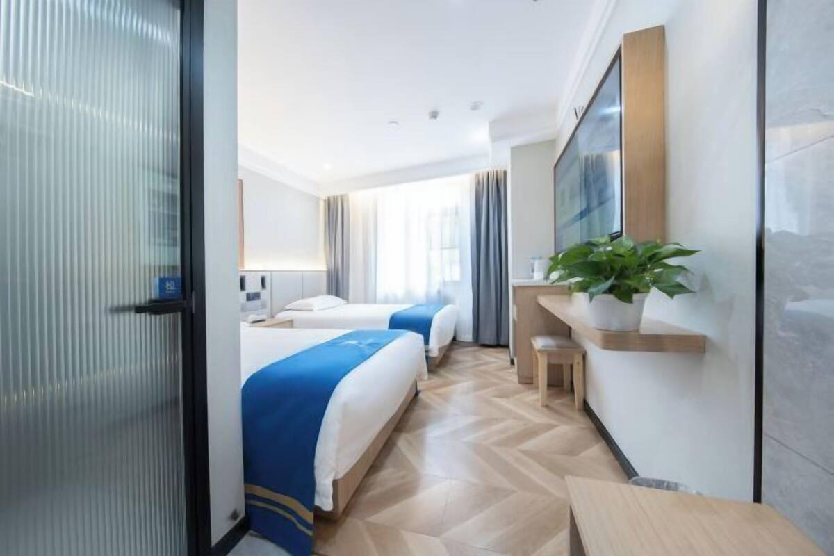 A hotel room with two beds, a desk, a chair, a plant, and a large window.