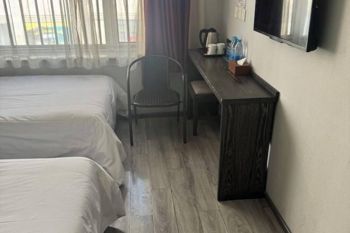A hotel room with two beds, a wooden desk, a chair, a television, and a window with sheer curtains.
