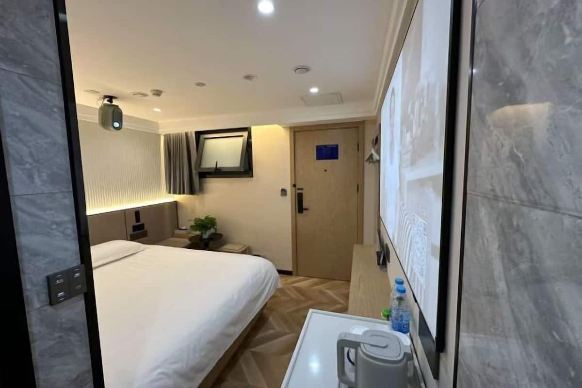 A hotel room with a bed, bedside table with a kettle, a nightstand, a lamp, a mirror, a door, and a wall-mounted TV.