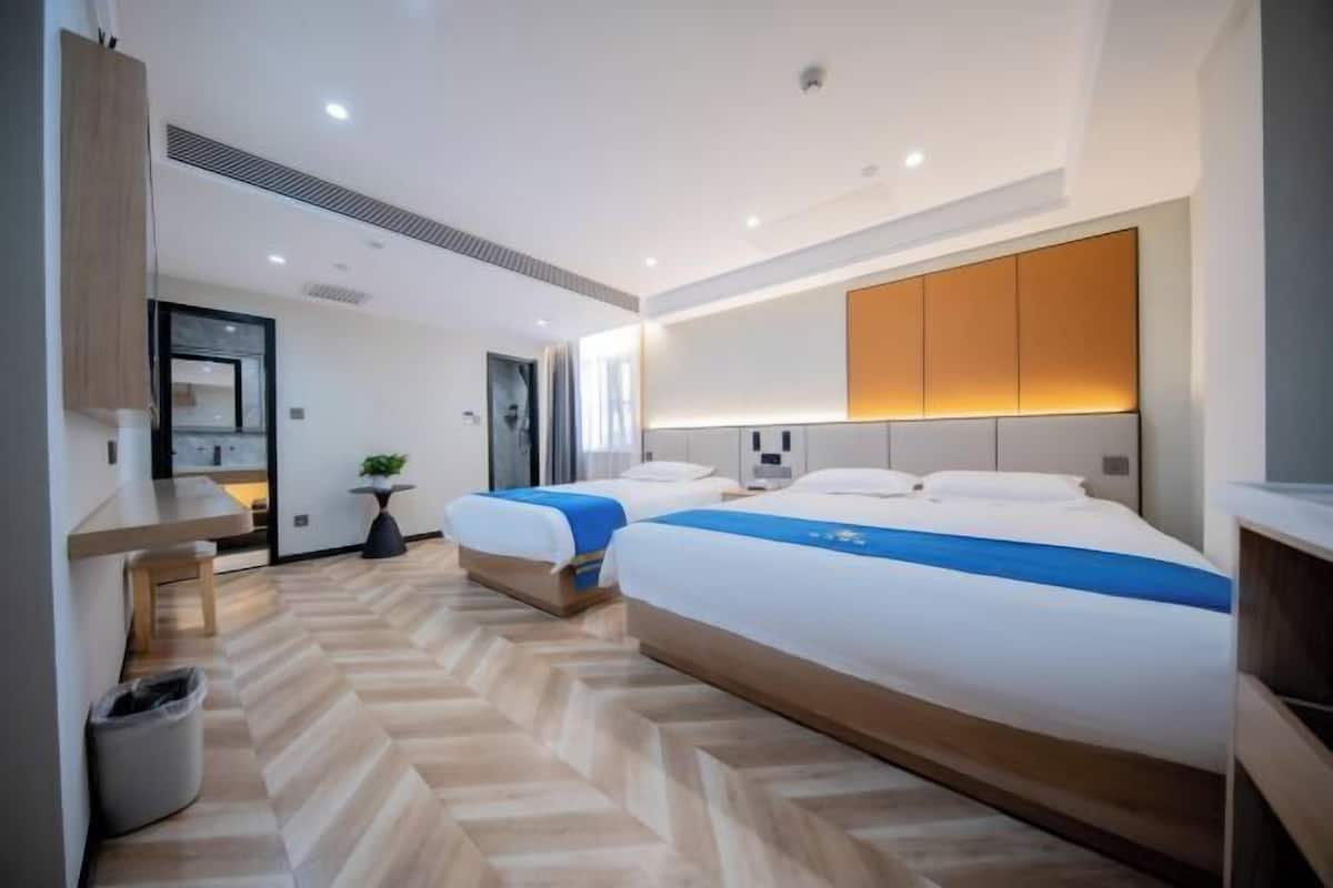A hotel room with two beds, a wooden herringbone floor, a bedside table, a trash bin, and a wall-mounted cabinet.