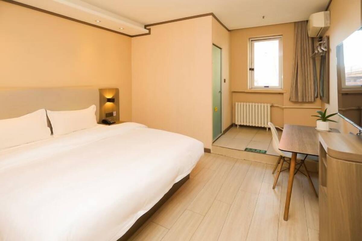 Habitación de hotel con cama, escritorio, silla, radiador, ventana y puerta.