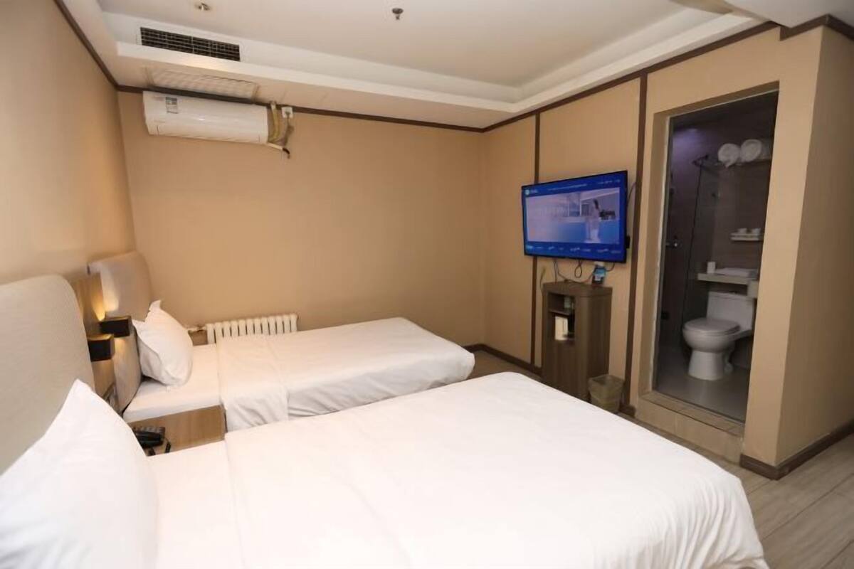 Habitación de hotel con dos camas individuales, televisión, baño con cabina de ducha de vidrio y equipo de aire acondicionado.