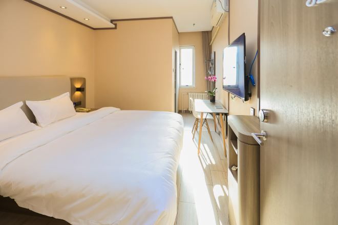 A hotel room with a large bed, a bedside table with a lamp, a TV mounted on the wall, and a window providing natural light.