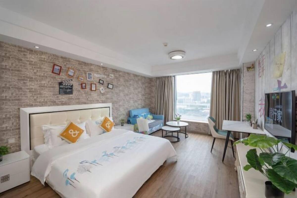 A hotel room with a brick wall, a bed with a white headboard, a blue sofa, a small round table, and a TV on the wall.
