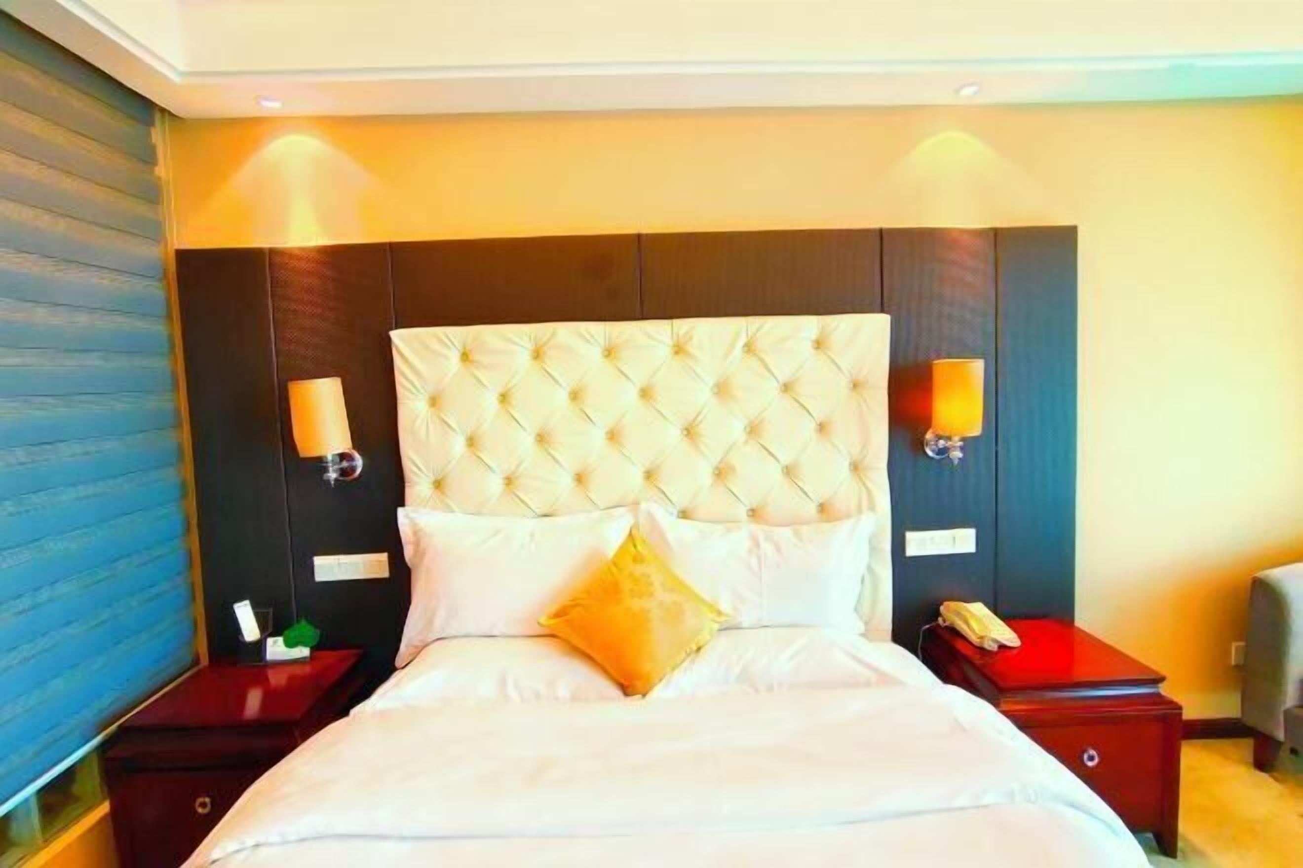 Una habitación de hotel con cabecera capitonada, dos lámparas de noche, una cama con ropa blanca y una mesita de noche con teléfono.