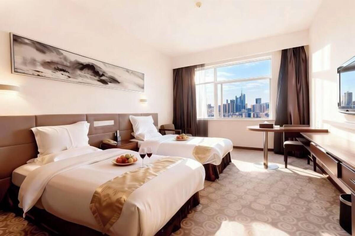 A hotel room with two beds, a desk, a large window with a city view, and a painting on the wall.