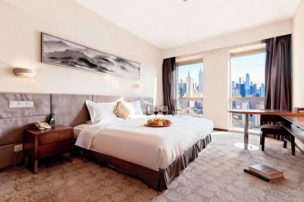 A hotel room with a large bed, a desk, a city view, and a painting on the wall.