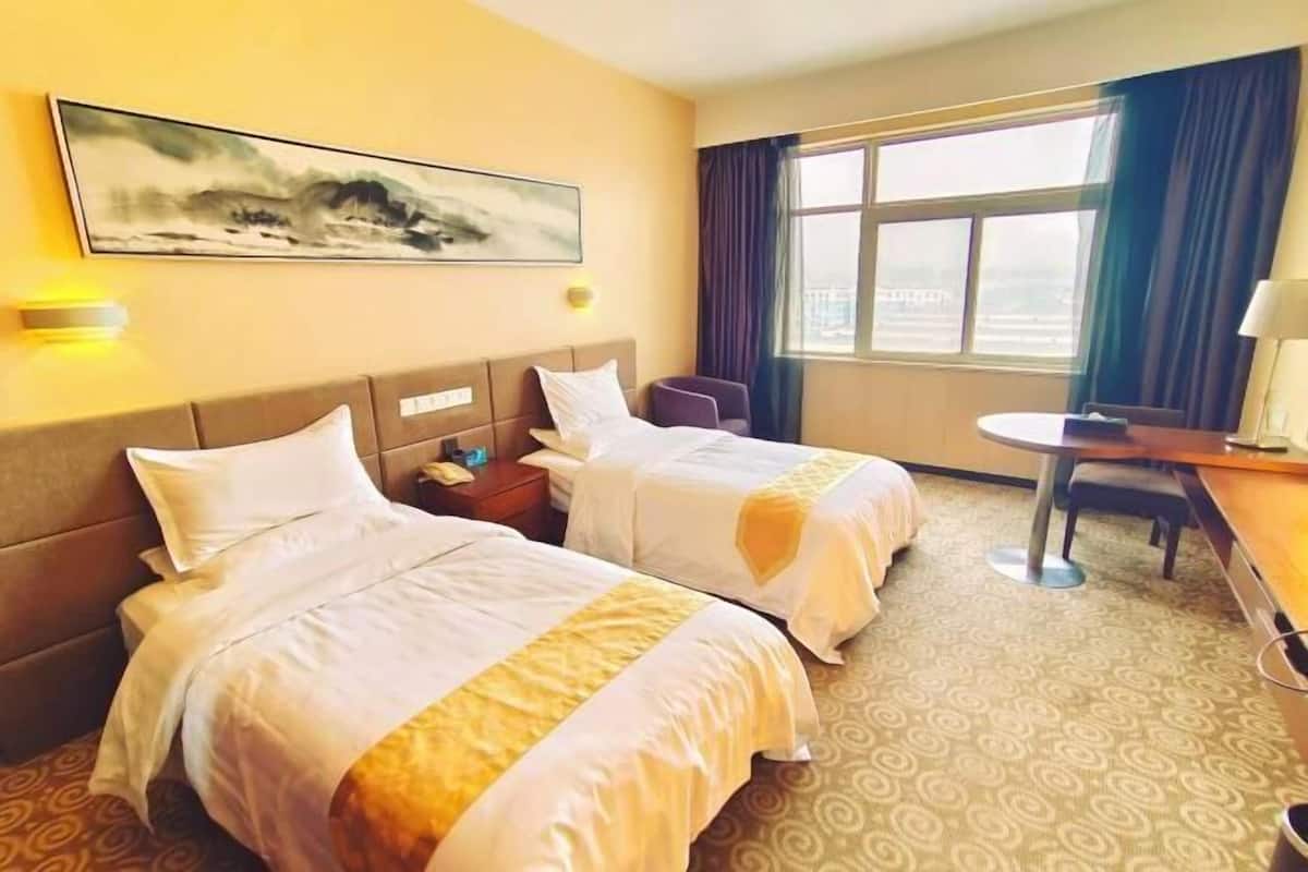 A hotel room with two beds, a small table, a chair, a painting on the wall, and a window with curtains.
