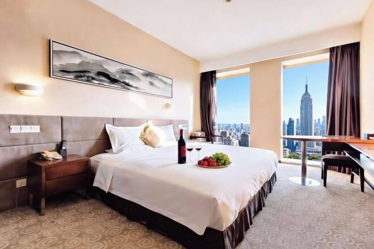 A hotel room with a bed, a desk, a chair, a city view, and a painting on the wall.