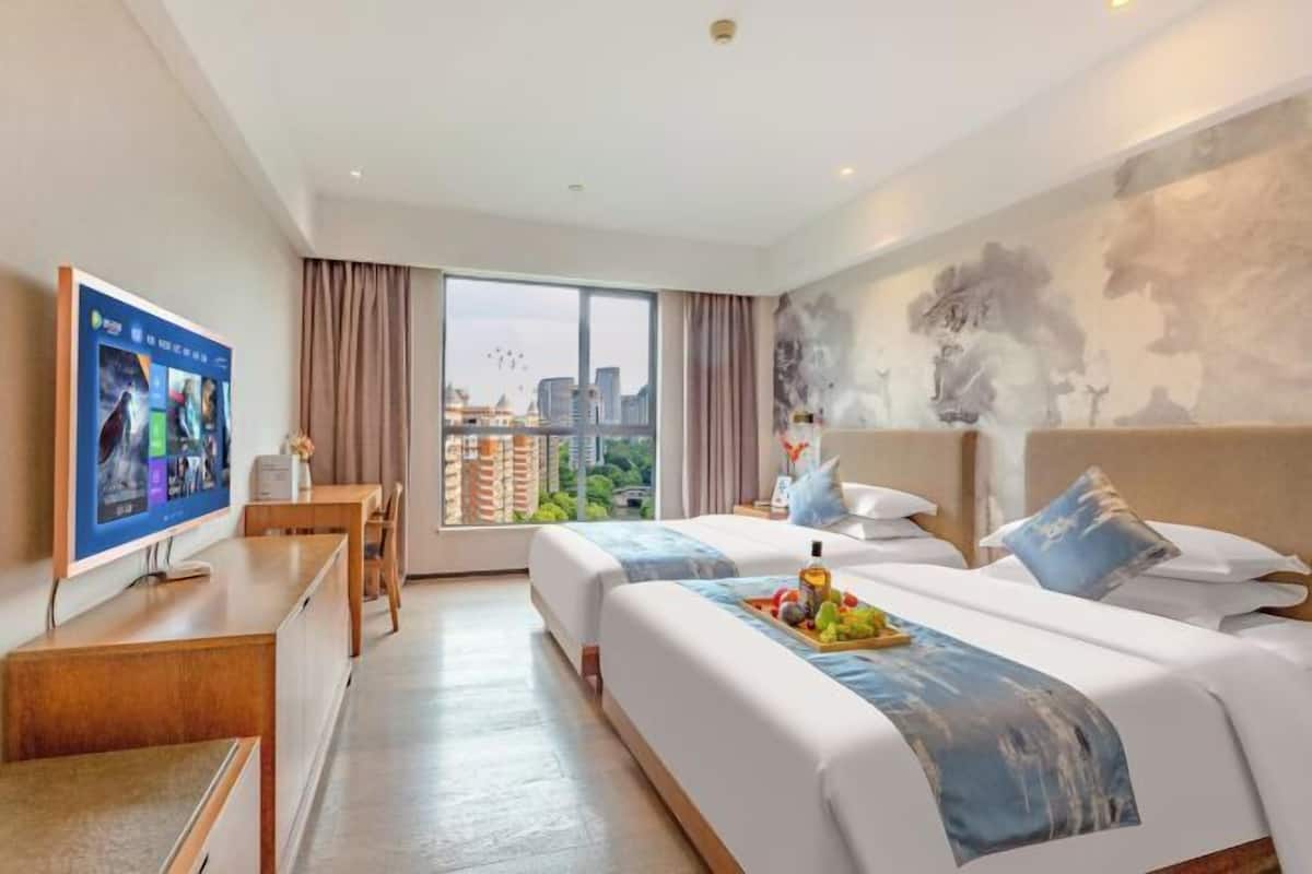 A hotel room with two beds, a TV, a desk, and a large window with a city view.